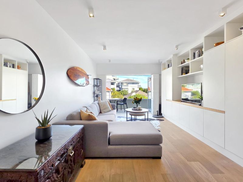 A bright and modern living room featuring custom built-in cabinetry, hardwood flooring, and bi-fold doors leading to a balcony with neighborhood views.