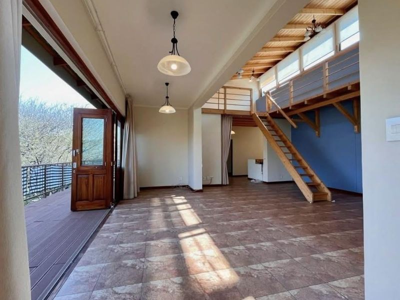 Spacious open-plan living area featuring a timber staircase leading to a mezzanine level, tiled flooring, and direct access to an outdoor balcony.