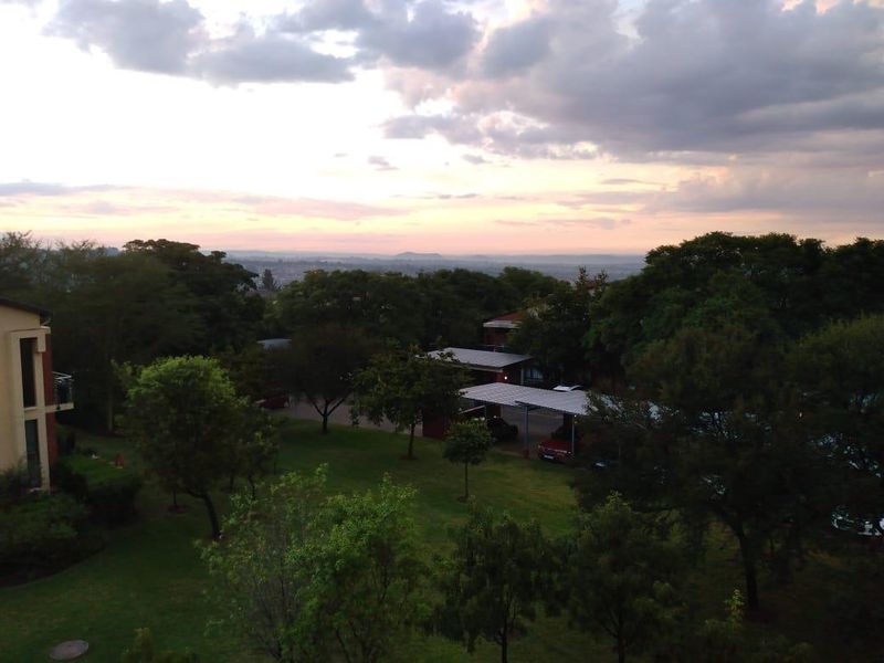 Elevated scenic view overlooking the apartment complex grounds, featuring mature trees, landscaped lawns, and covered parking areas at sunset.