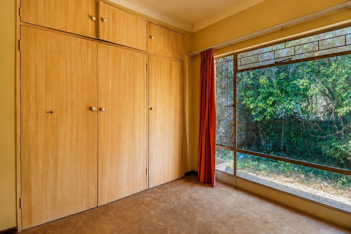 A vacant bedroom featuring extensive built-in wooden cabinetry, large windows with garden views, and carpeted flooring, offering a blank canvas for renovation.