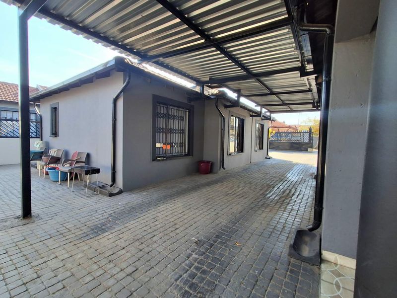 A paved rear exterior area featuring a covered carport structure, security bars on windows, and functional rainwater drainage systems.
