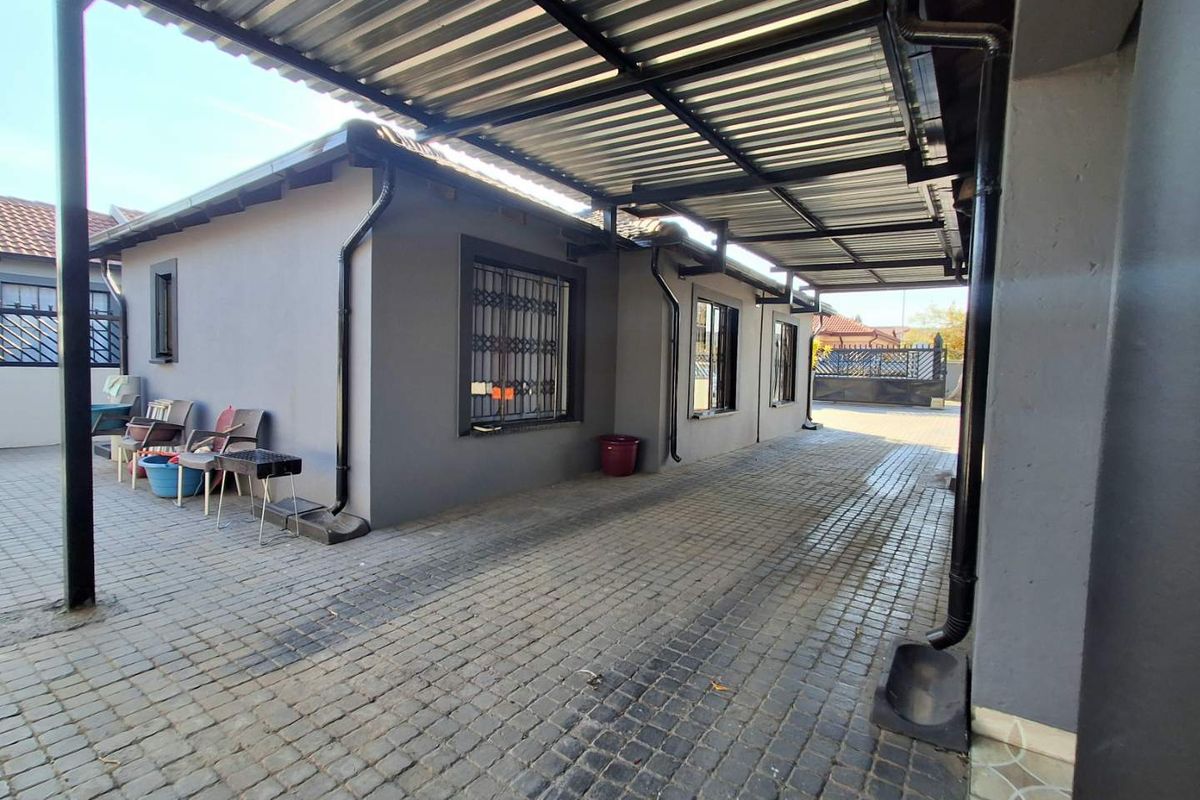 A paved rear exterior area featuring a covered carport structure, security bars on windows, and functional rainwater drainage systems.