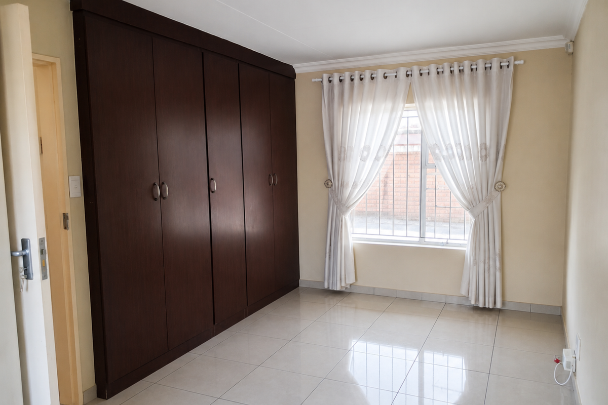 A clean, empty bedroom featuring large dark wood built-in cupboards, neutral tiled flooring, and a window fitted with security bars.