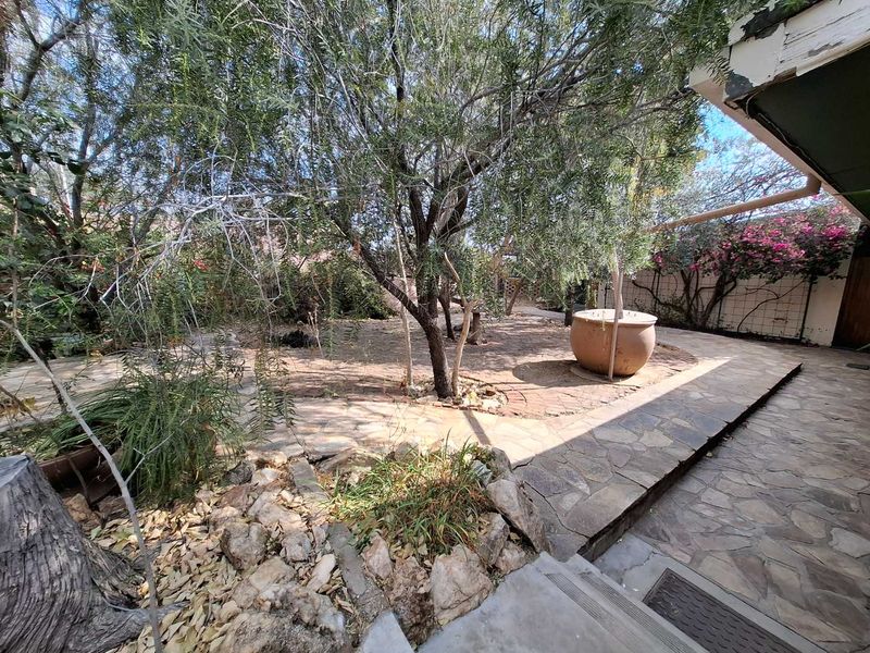 A rear garden area featuring a stone-paved patio, mature trees, and a trellis, requiring maintenance to the roof eaves and landscaping.