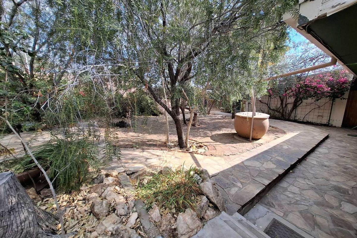 A rear garden area featuring a stone-paved patio, mature trees, and a trellis, requiring maintenance to the roof eaves and landscaping.