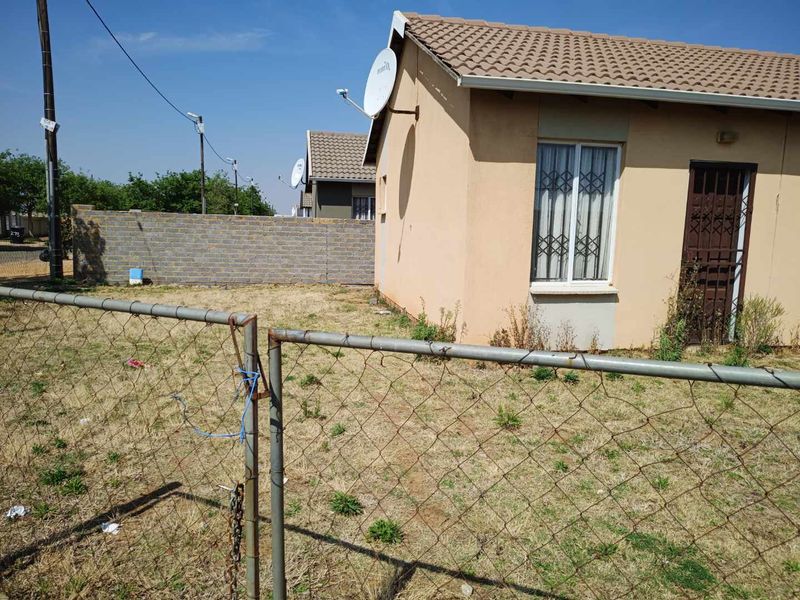 Exterior view of a single-story house featuring a tiled roof, security gate, and a large, unmaintained garden area enclosed by a chain-link fence.