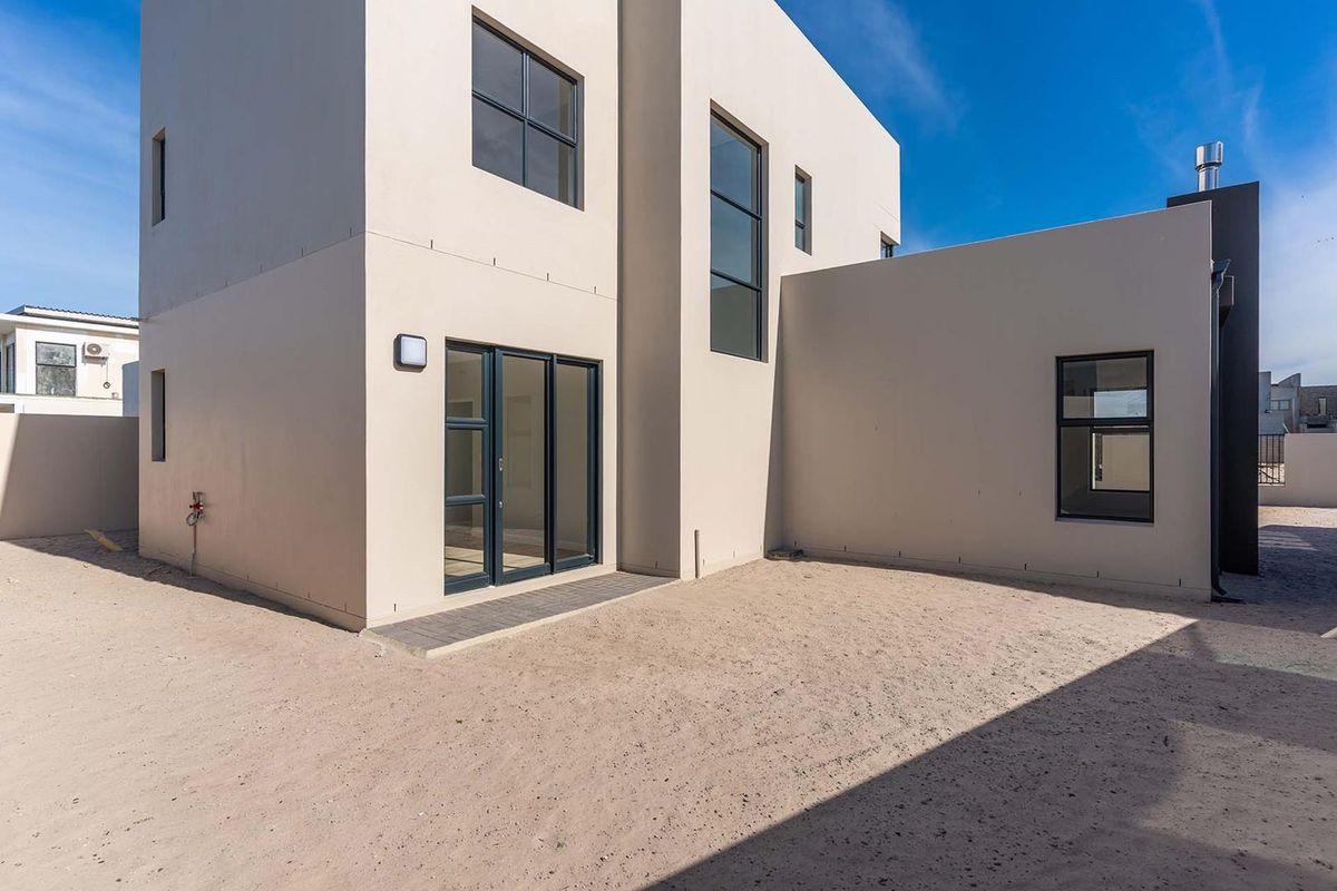 Rear view of a modern double-storey house featuring clean rendered walls, large aluminium-framed windows, and a spacious, undeveloped sandy backyard area.