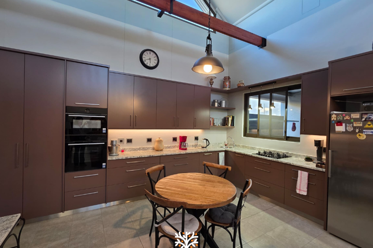 Modern kitchen featuring dark cabinetry, stone countertops, built-in double oven, gas hob, and high vaulted ceilings with exposed timber beams.