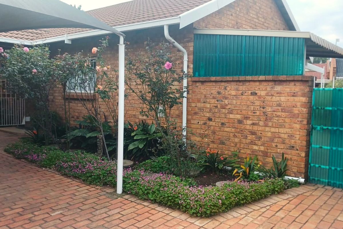 A rear exterior view of a face brick cluster home featuring a paved patio area, shade sail, and established garden beds.