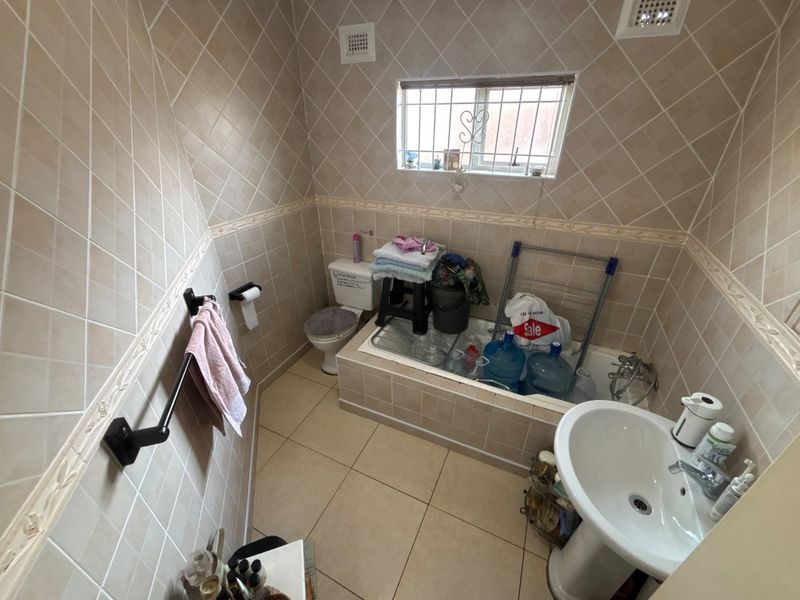 A functional bathroom featuring a pedestal basin, toilet, and built-in bathtub with full-height wall tiling and a window with security bars.