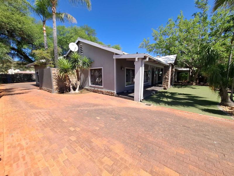 Front exterior view of the house featuring a brick paved driveway, covered patio area, and well-maintained artificial lawn with stone cladding accents.