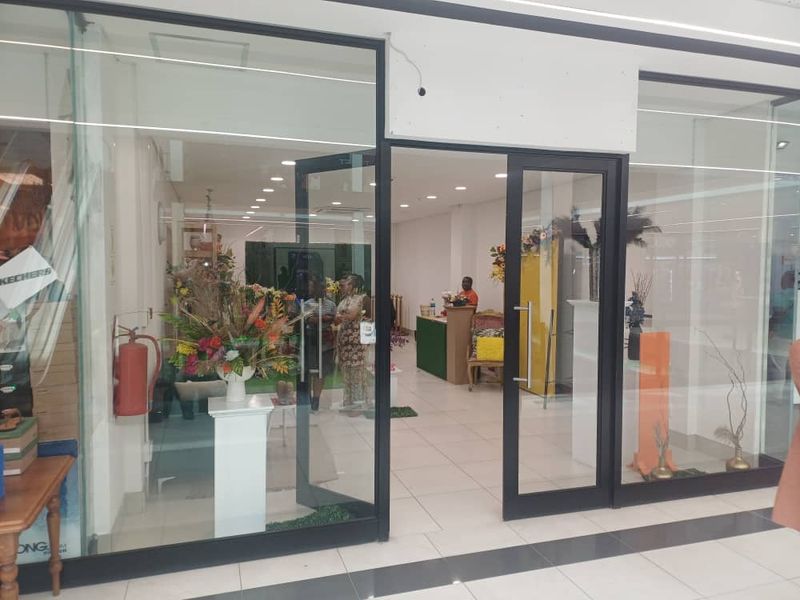 Retail storefront entrance featuring a glass facade with black aluminum framing and recessed ceiling lighting, located within a shopping center.