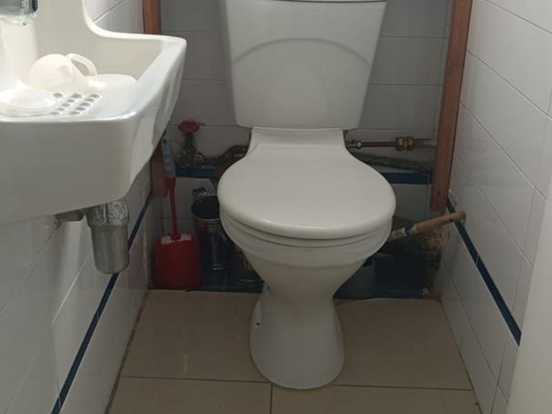A compact bathroom featuring a wall-mounted basin and toilet, with tiled finishes and exposed plumbing requiring cosmetic attention.