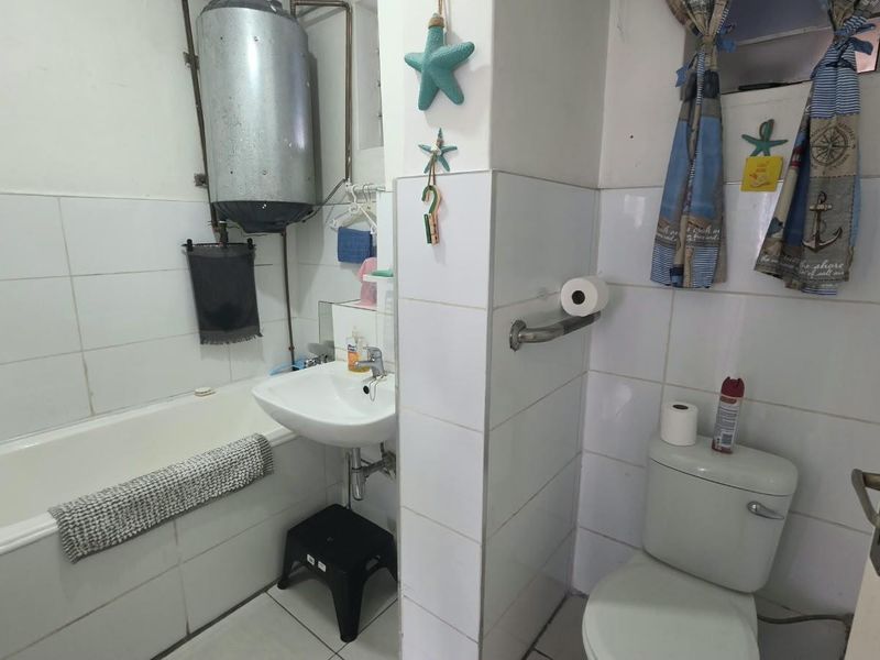 A functional bathroom featuring a bathtub, pedestal basin, and toilet, with tiled finishes and an exposed wall-mounted geyser unit.