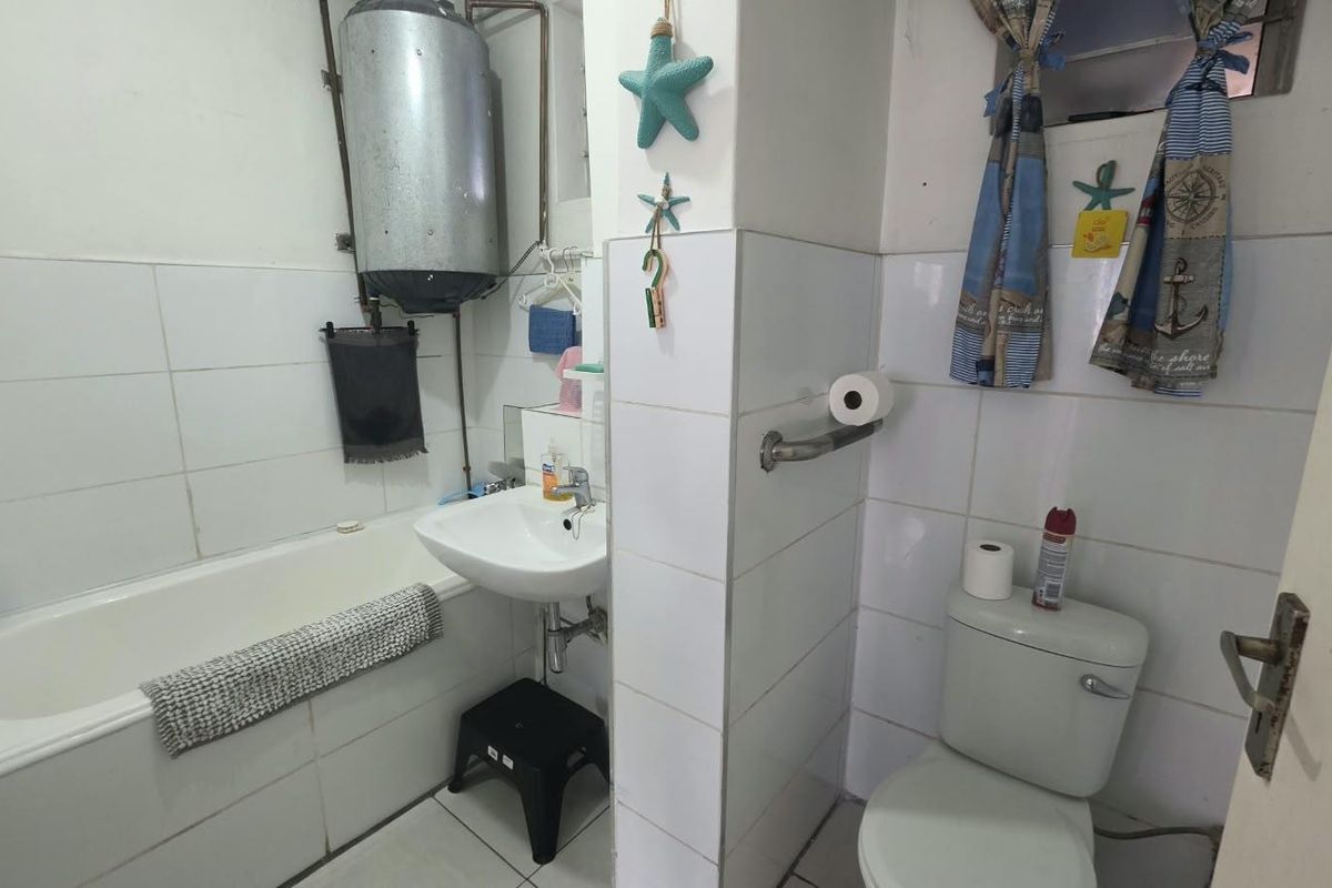 A functional bathroom featuring a bathtub, pedestal basin, and toilet, with tiled finishes and an exposed wall-mounted geyser unit.