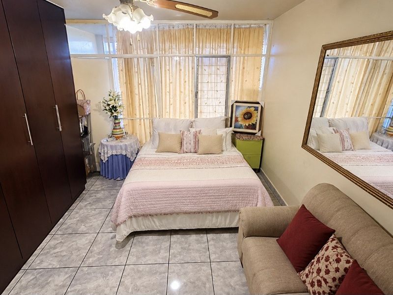 A functional bedroom featuring tiled flooring, a large built-in wardrobe, and a ceiling fan, with natural light provided by a large window.