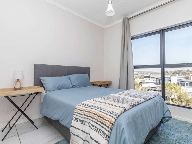 A bright bedroom featuring large aluminium-framed windows that provide natural light and expansive views, complemented by neutral tiled flooring.
