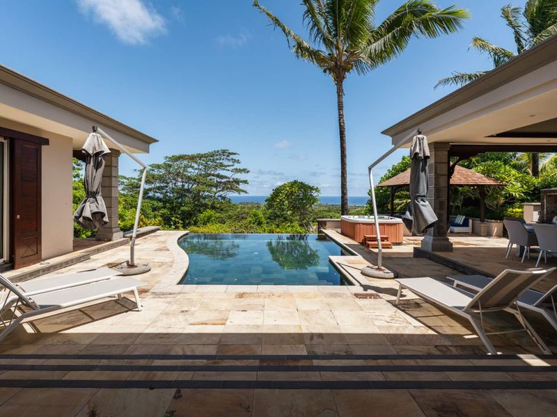 A luxurious outdoor pool area featuring a tiled deck, infinity-style pool, integrated spa, and covered patio space with scenic ocean views.