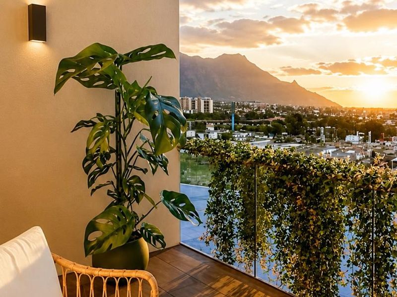 A modern balcony featuring timber-look decking, a glass balustrade with integrated greenery, and a wall-mounted light fixture, offering scenic sunset mountain views.