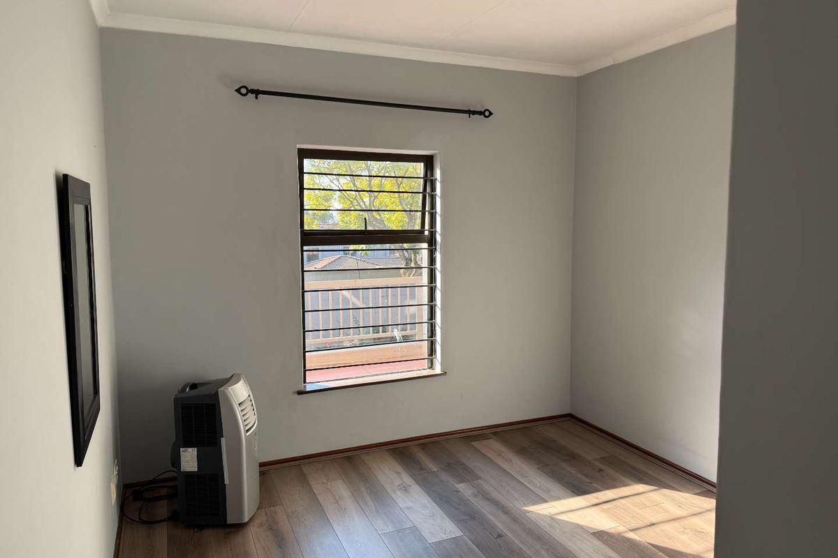 A bright, empty bedroom featuring modern laminate flooring, neutral grey walls, and a window fitted with security bars.