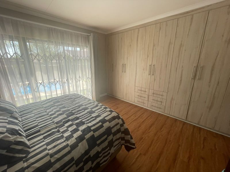 A well-maintained bedroom featuring extensive built-in cupboards, warm laminate flooring, and a sliding door providing access to the outdoor pool area.