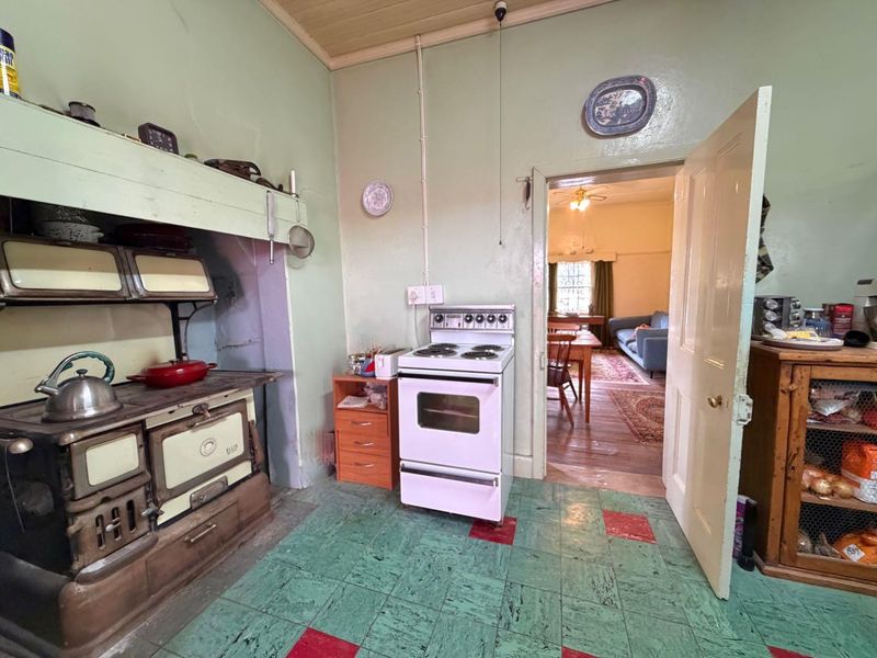 A dated kitchen featuring a vintage cast iron stove, freestanding electric oven, timber ceiling, and patterned vinyl flooring, with an open doorway to the living area.