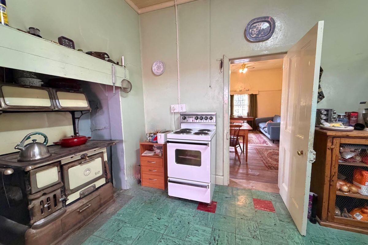 A dated kitchen featuring a vintage cast iron stove, freestanding electric oven, timber ceiling, and patterned vinyl flooring, with an open doorway to the living area.