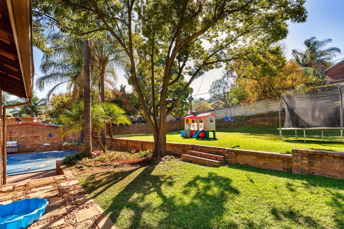 A spacious rear garden featuring a terraced lawn, mature trees, a brick retaining wall, and a swimming pool with a cover.