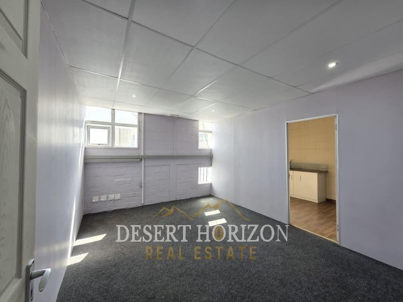 An empty office space featuring carpet flooring, a suspended ceiling with recessed lighting, and painted masonry walls, with an adjacent kitchenette area.