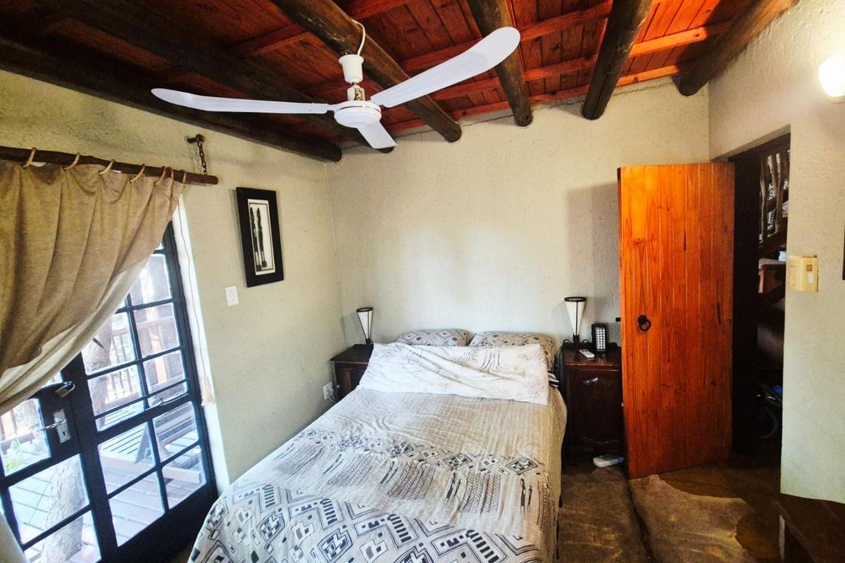 A rustic bedroom featuring exposed timber ceiling beams, a ceiling fan, and a glass-paneled door providing access to an outdoor area.