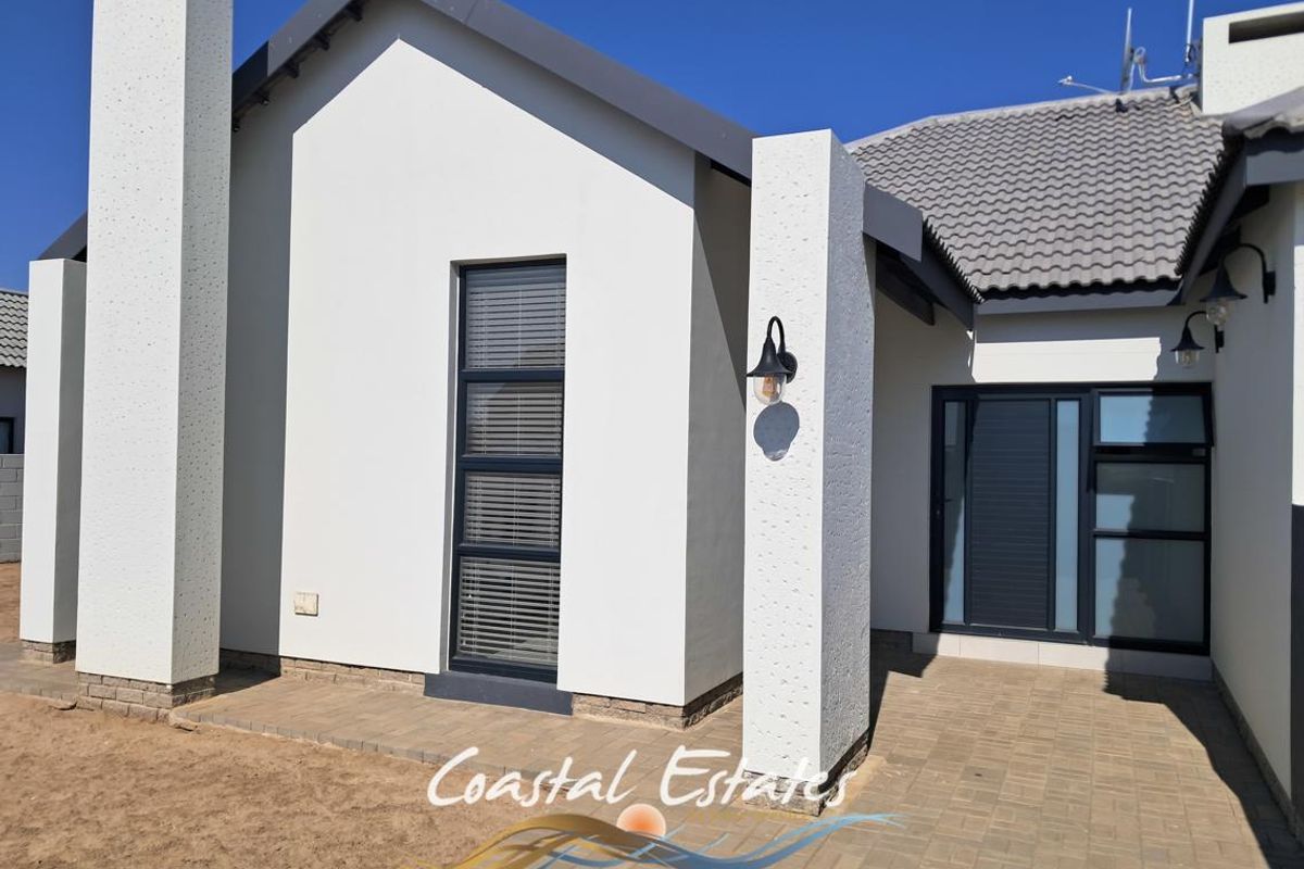 A modern rear exterior view featuring a paved patio area, clean white rendered walls, tiled roofing, and contemporary aluminium window and door frames.