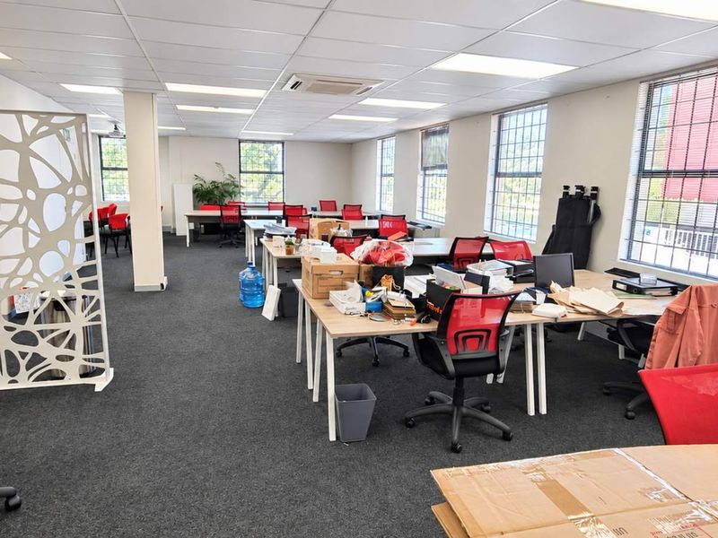 Spacious open-plan office area featuring carpeted flooring, suspended ceilings with recessed lighting, and large windows providing ample natural light.