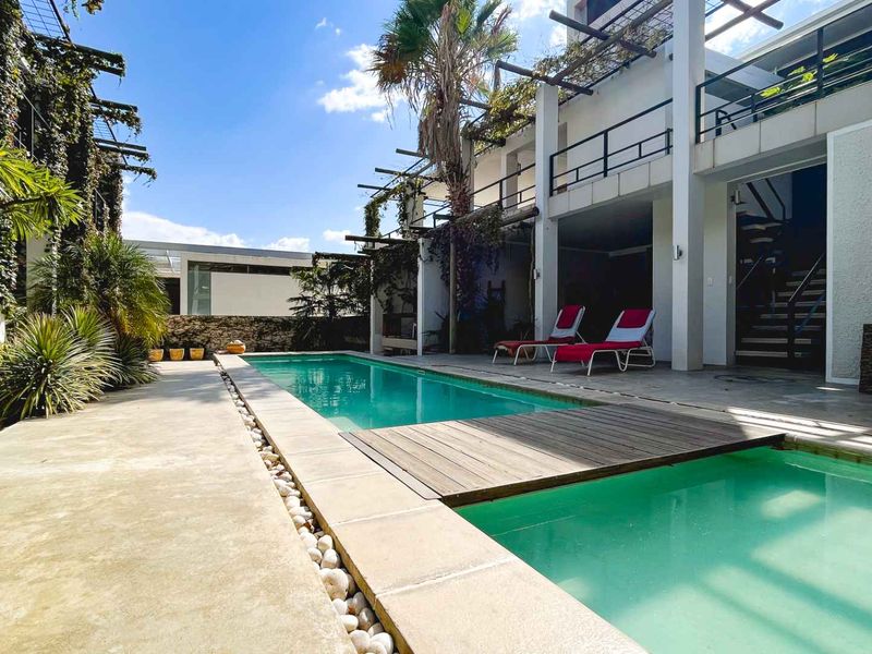 A well-maintained outdoor pool area featuring a swimming pool with a wooden deck, stone paving, and a covered patio with access to upper levels.