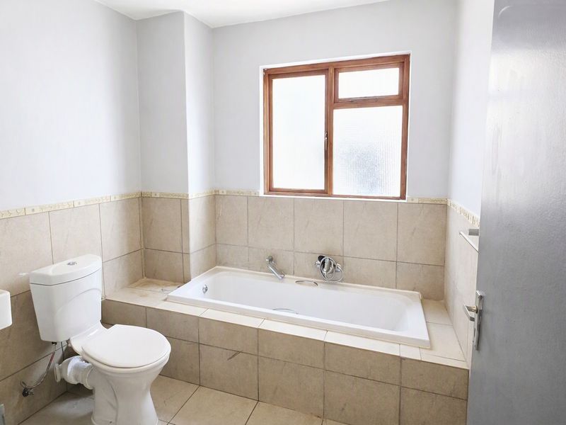 A functional bathroom featuring a built-in bathtub, toilet, and neutral tiled finishes with a timber-framed window.