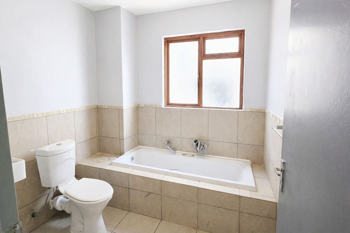 A functional bathroom featuring a built-in bathtub, toilet, and neutral tiled finishes with a timber-framed window.
