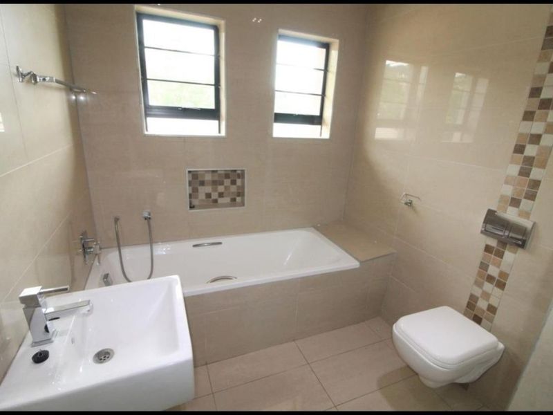 Modern bathroom featuring a built-in bathtub, wall-mounted toilet, basin, and neutral tiled finishes with a decorative mosaic accent.