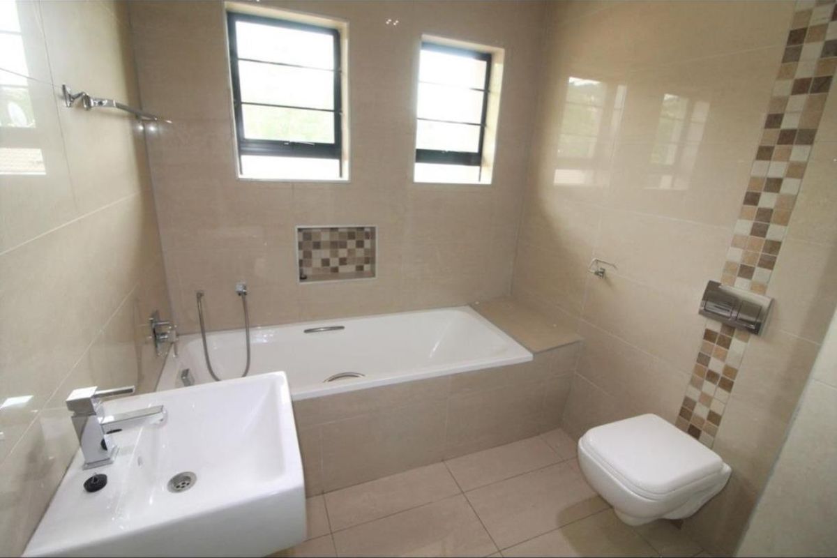 Modern bathroom featuring a built-in bathtub, wall-mounted toilet, basin, and neutral tiled finishes with a decorative mosaic accent.