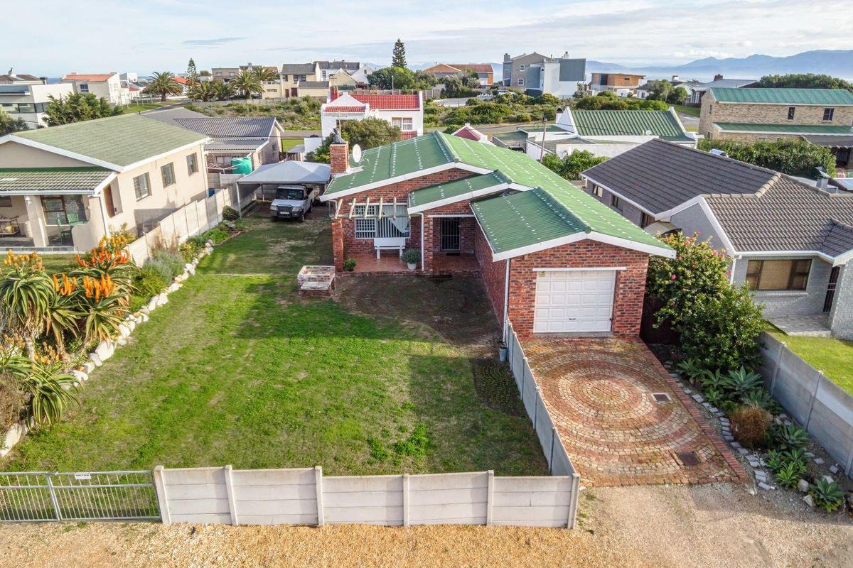 Aerial view of a single-story brick house featuring a green metal roof, paved driveway, attached garage, and a spacious front garden with boundary walling.