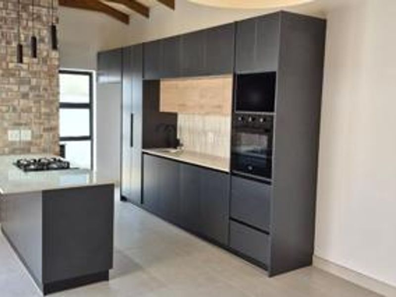 Modern kitchen featuring dark cabinetry, stone countertops, a gas hob on the island, and an exposed brick wall with timber ceiling beams.