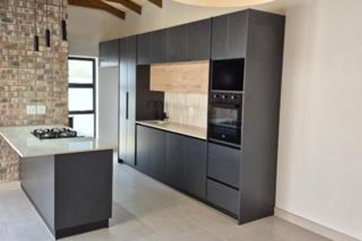 Modern kitchen featuring dark cabinetry, stone countertops, a gas hob on the island, and an exposed brick wall with timber ceiling beams.