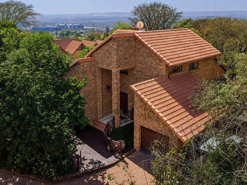 Aerial view of a multi-level brick house with a terracotta tile roof, featuring a paved driveway, garage, and wooden deck set in a lush environment.
