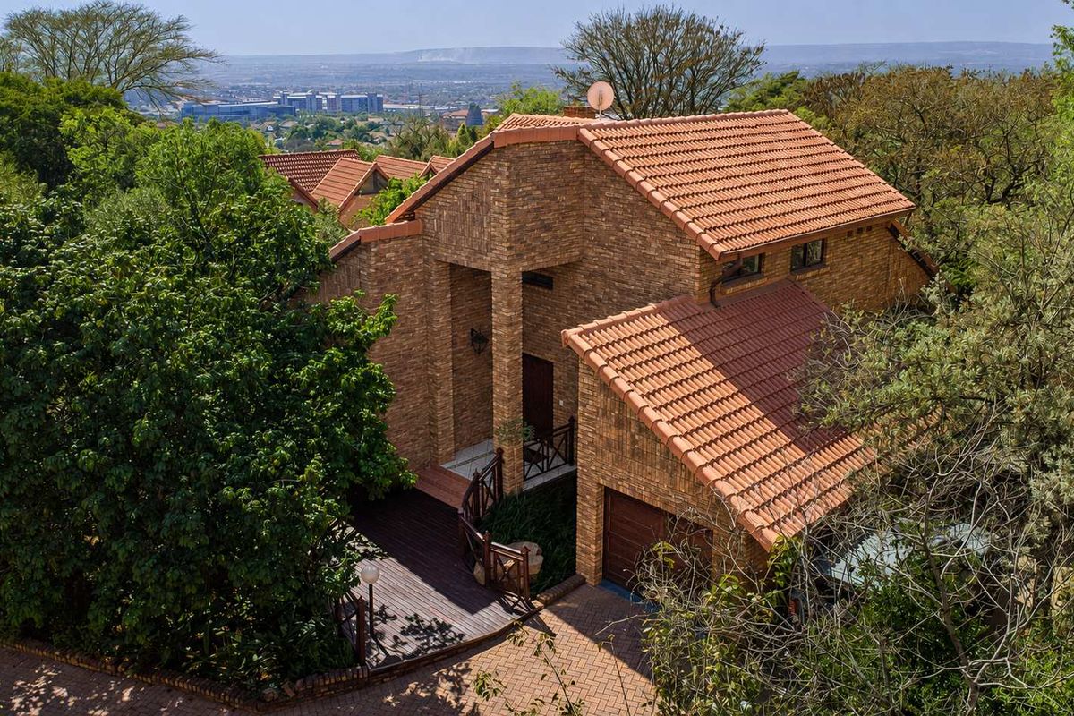 Aerial view of a multi-level brick house with a terracotta tile roof, featuring a paved driveway, garage, and wooden deck set in a lush environment.