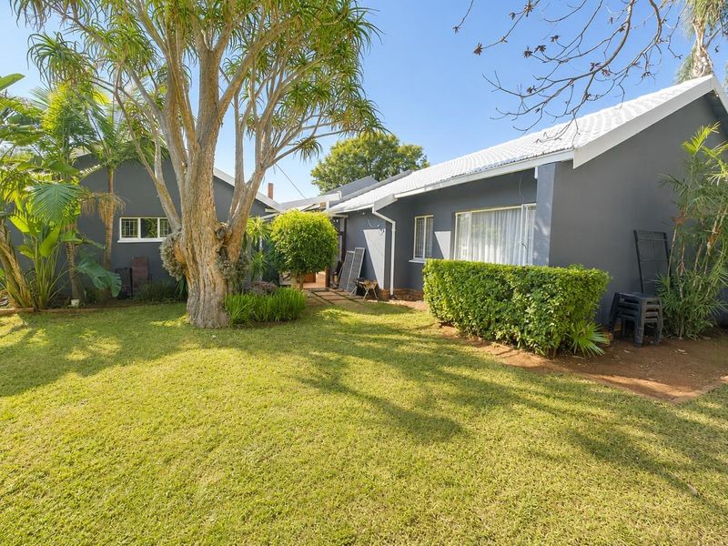 A well-maintained rear exterior view of the house featuring a spacious lawn, mature trees, and a paved pathway leading to the building entrance.