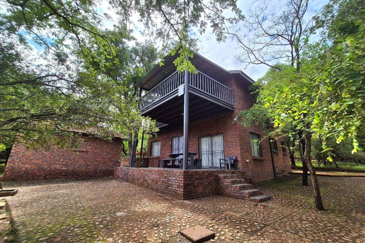 A two-story brick house featuring a covered ground-floor patio and an upper-level balcony, set within a mature, tree-lined garden with paved courtyard.