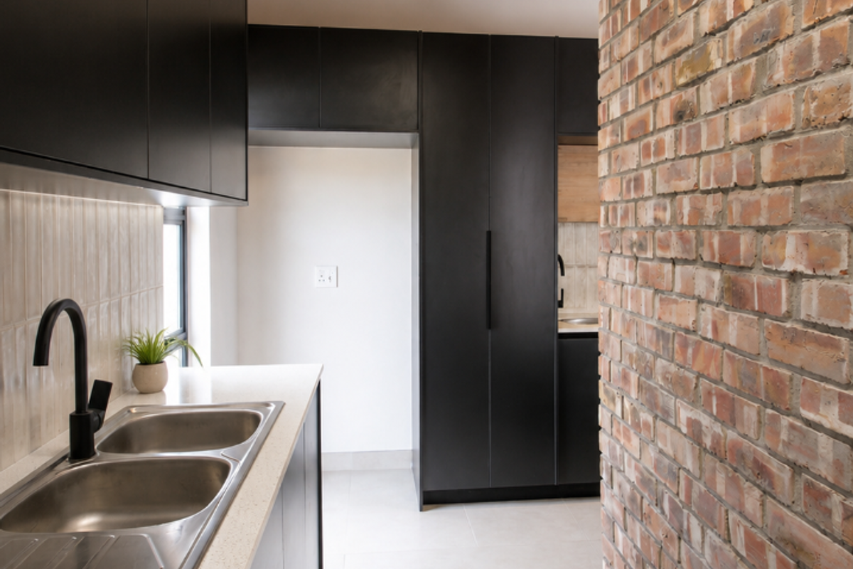 Modern kitchen featuring matte black cabinetry, stone countertops, a double sink, and a striking exposed brick wall under a timber-beamed ceiling.