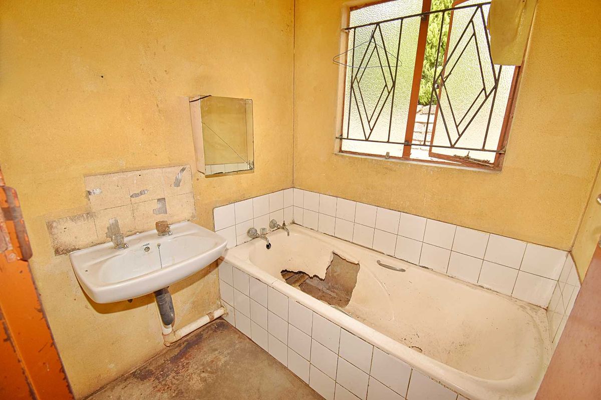 A dilapidated bathroom featuring a damaged bathtub, wall-mounted basin, and window with security bars, requiring significant renovation and repair.