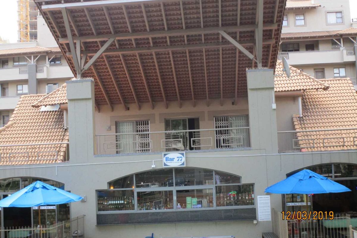 Exterior view of a multi-level apartment building featuring a covered balcony, tiled roofing, and ground-level commercial space with large windows.