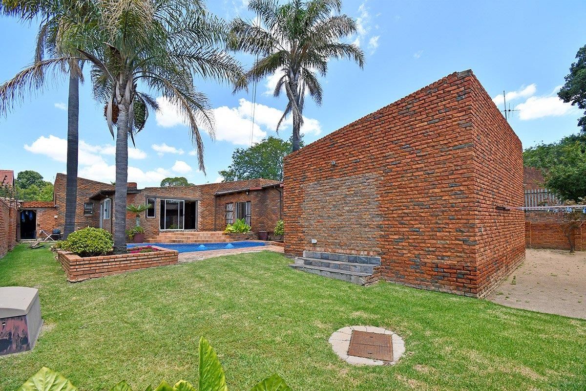 Rear garden area featuring a swimming pool, established palm trees, and a prominent face-brick exterior structure with a lawned yard.