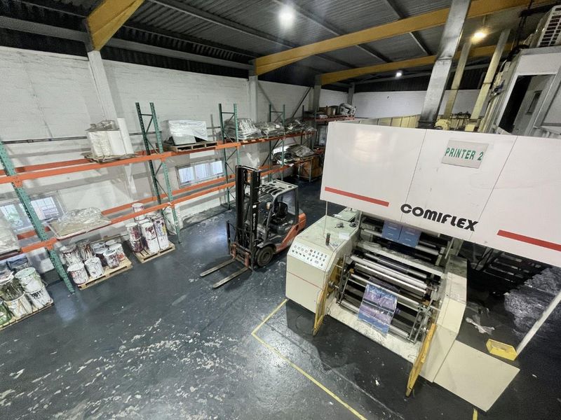 Industrial warehouse space featuring high ceilings with exposed steel trusses, heavy-duty racking systems, and integrated industrial printing equipment on a durable concrete floor.