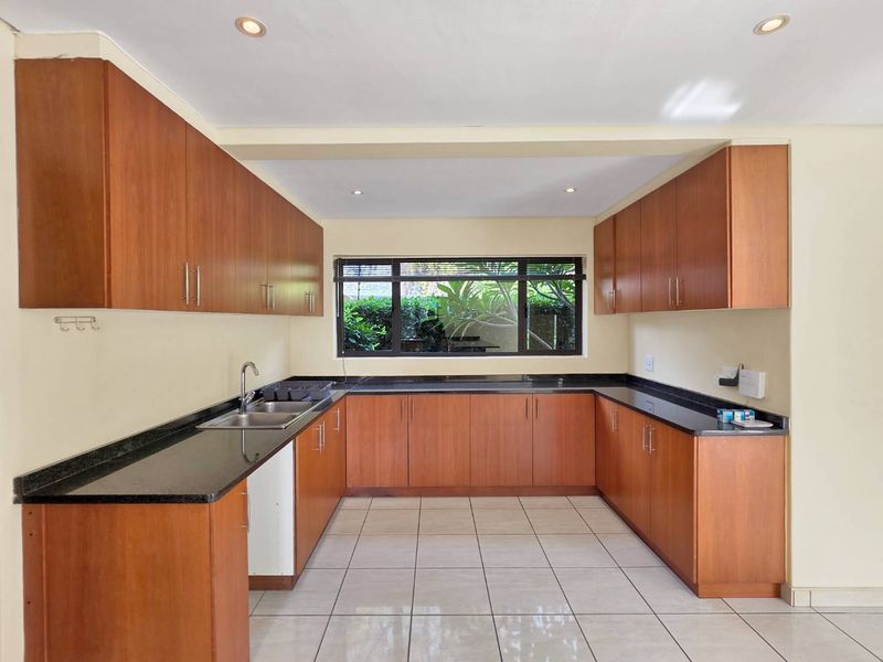 A clean, U-shaped kitchen featuring warm wood-toned cabinetry, dark granite countertops, tiled flooring, and a large window providing natural light.